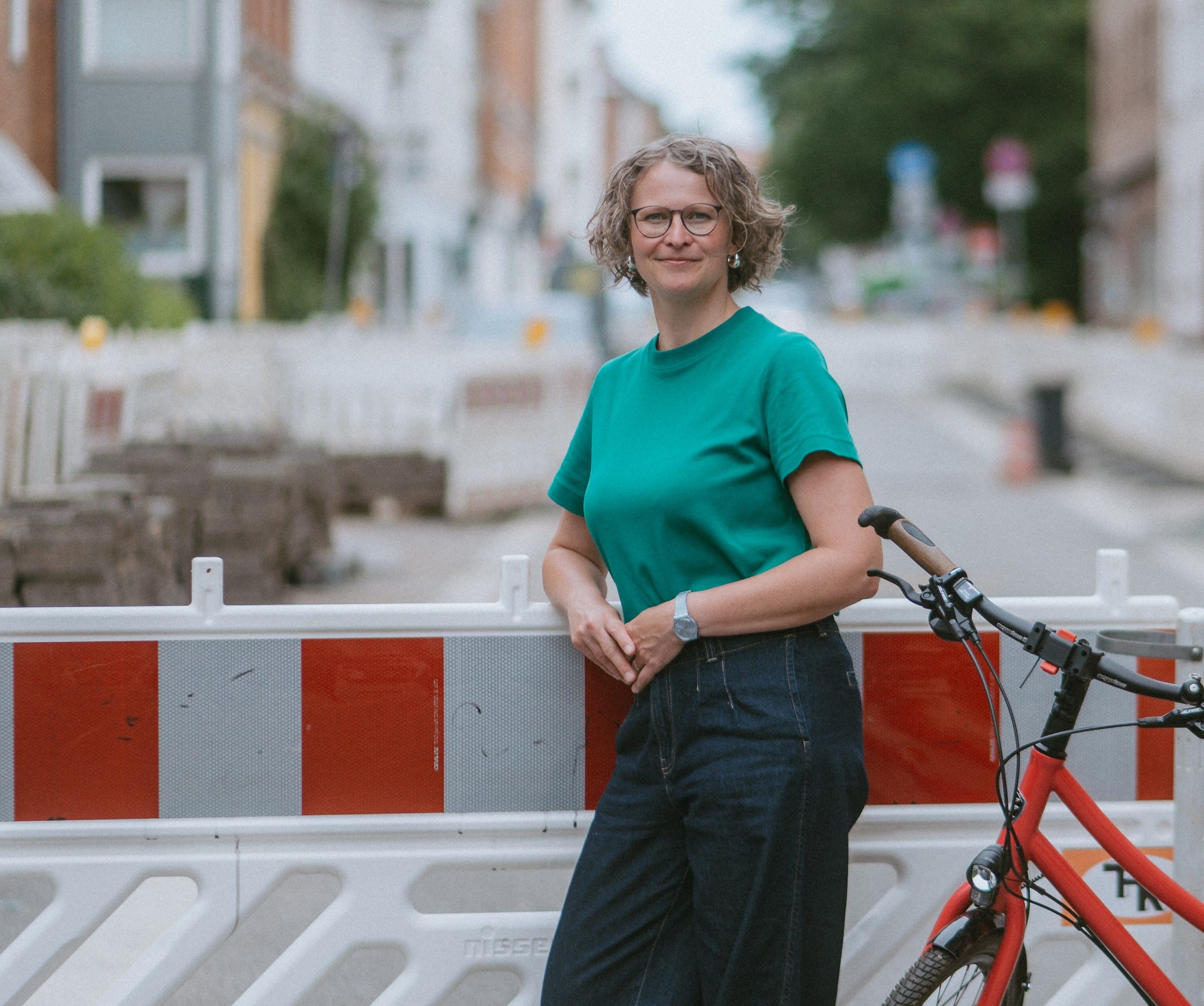 Eva lehnt an einer rot-weißen Baustellenabsperrung in einer Kieler Straße, neben ihr ihr rotes Fahrrad