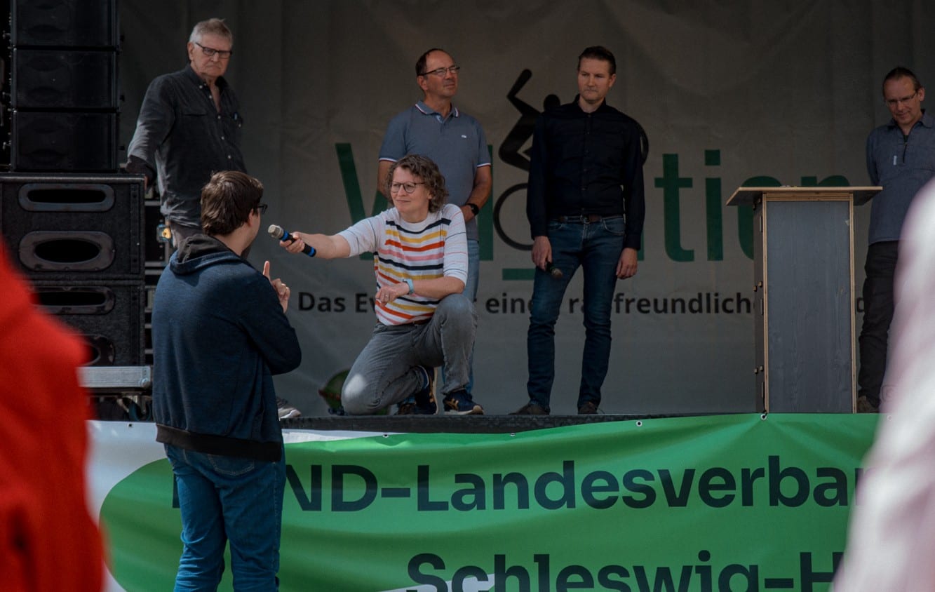 Eva kniet am Bühnenrand und reicht einem Besucher das Mikrofon, hinter ihr die Gäste auf der Bühne
